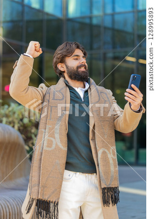 Happy Indian man using smartphone celebrating victory win good message news on downtown city street 128995893