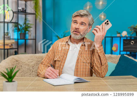 Mature man at sofa table on mobile phone speaker call, writing notes in notebook, serious at home Mature man at sofa table on mobile phone speaker call, writing notes in notebook, serious at home 128995899