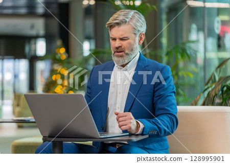 Sad businessman using laptop loses becoming shocked sudden lottery results in modern hotel lobby Sad businessman using laptop loses becoming shocked sudden lottery results in modern hotel lobby 128995901