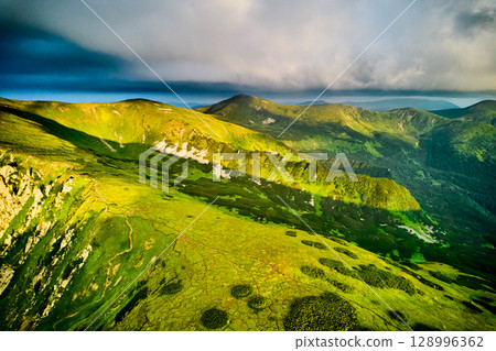 Breathtaking aerial view of green, rugged mountain peaks under dramatic sky. Scene captures contrast between vibrant landscape and moody clouds, with sunlight breaking through. Carpathians, Chornogora Breathtaking aerial view of green, rugged mountain peaks under dramatic sky. Scene captures contrast between vibrant landscape and moody clouds, with sunlight breaking through. Carpathians, Chornogora 128996362
