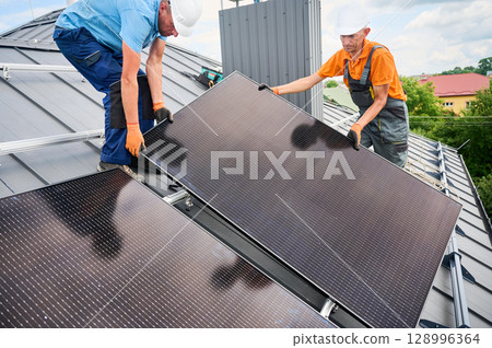 Workers building solar panel system on rooftop of house. Two men installers in helmets installing photovoltaic solar module outdoors. Alternative, green and renewable energy generation concept. 128996364