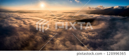 Aerial view of vibrant sunset over white dense clouds bathes in warm golden light, creating dreamlike landscape. Soft shadows play across undulating clouds, while distant landforms peek through mist. 128996365