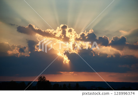 Sun sets behind dramatic clouds, casting rays of golden light. Sky painted with warm hues of orange, pink, contrasting with darker clouds. Silhouette trees, distant houses add depth to evening scene. Sun sets behind dramatic clouds, casting rays of golden light. Sky painted with warm hues of orange, pink, contrasting with darker clouds. Silhouette trees, distant houses add depth to evening scene. 128996374