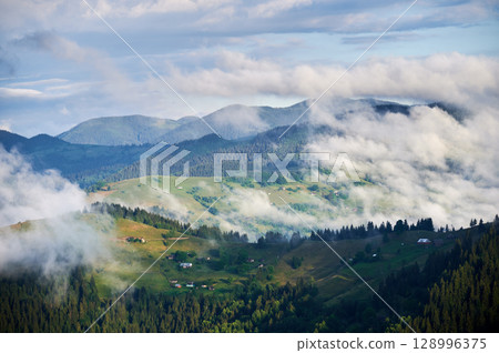 Picturesque scene of mountain valley blanketed in mist, with patches of green pastures and scattered houses. Clouds drift gently over forested hills, creating serene and tranquil atmosphere. 128996375