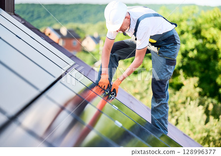 Worker building photovoltaic solar panel system on rooftop of house. Man engineer in helmets and gloves installing solar module with help of hex key outdoors. Renewable energy generation concept. 128996377