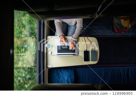 Woman working on laptop while traveling. Computer and smartphone connected to power bank, top view. 128996905