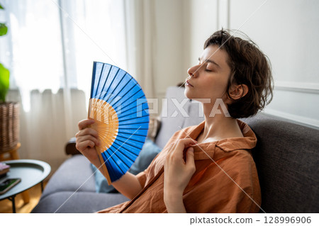 Exhausted sweaty woman fanning herself sitting on sofa suffering from summer heat without AC 128996906