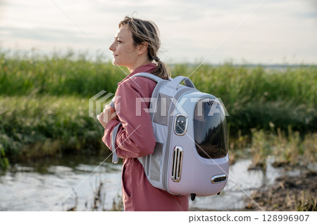 Carefree woman tourist walking riverside trail carrying cat in backpack. Independent travel with pet 128996907