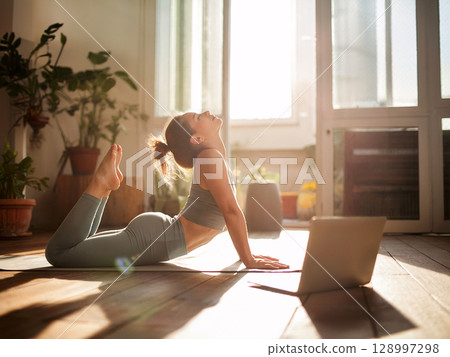 Young woman practicing bow pose during online yoga session at home surrounded by plants and natural sunlight Young woman practicing bow pose during online yoga session at home surrounded by plants and natural sunlight 128997298