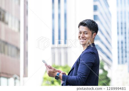 A young businessman operating a smartphone in an office district, looking at the camera 128999481