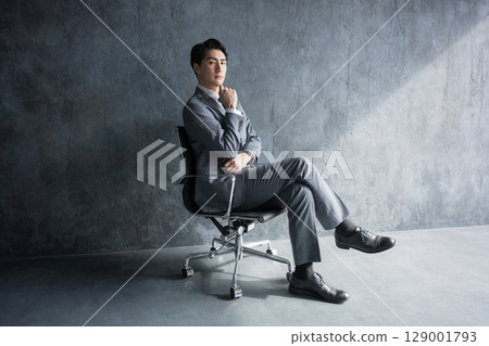一位西裝革履的商人全身坐在灰色背景的椅子上。一位經理或總裁思考戰略和戰術的形象 一位西裝革履的商人全身坐在灰色背景的椅子上。一位經理或總裁思考戰略和戰術的形象 129001793