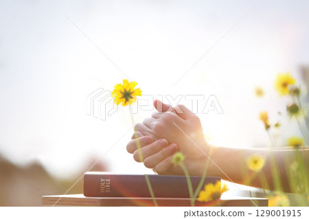 Christian praying with both hands together on the holy bible, beautiful sunny church with yellow flower field background Christian praying with both hands together on the holy bible, beautiful sunny church with yellow flower field background 129001915