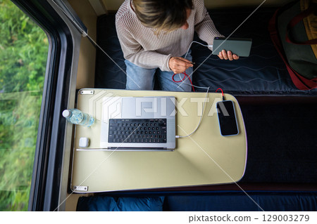 Woman plugging phone and laptop into power bank, charging batteries, sitting in train compartment. Woman plugging phone and laptop into power bank, charging batteries, sitting in train compartment. 129003279