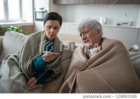 Puzzled shivering senior man and wife checking temperature on couch together. Fever ill symptoms 129003300