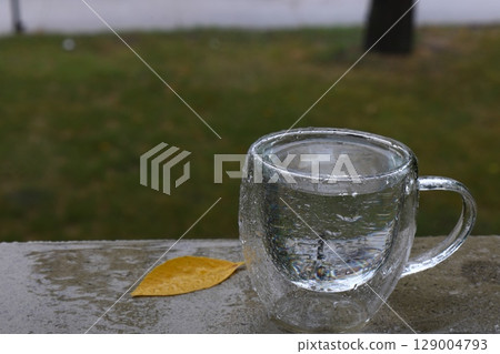 Glass Mug with Water and Yellow Leaf on Rainy Day  129004793