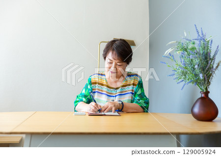 A senior woman in her 60s sitting at a table and writing in her notebook with a pen A senior woman in her 60s sitting at a table and writing in her notebook with a pen 129005924