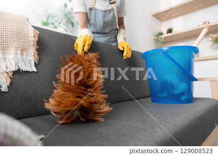 Home Cleaning. Woman dusting sofa with duster and bucket in modern living room. Home Cleaning. Woman dusting sofa with duster and bucket in modern living room. 129008523