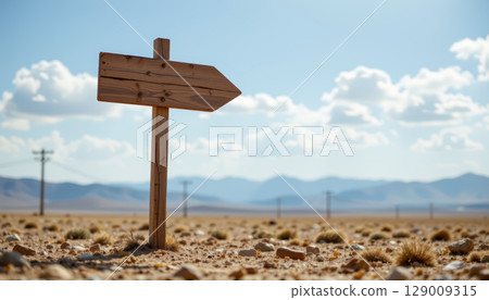 Wooden arrow signpost in dry desert landscape with rocky ground and distant mountains under blue sky 129009315