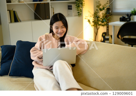 Smiling young woman relaxing on a sofa while using a laptop, enjoying online activities 129009601
