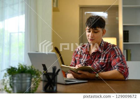 Freelancer in casual outfit writing ideas in journal next to laptop in a bright home workspace 129009672