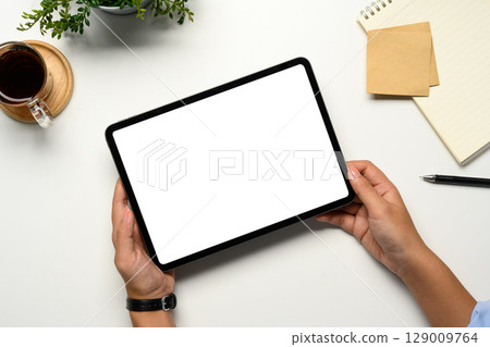 Hands holding a digital tablet with a blank white screen on a clean desk 129009764