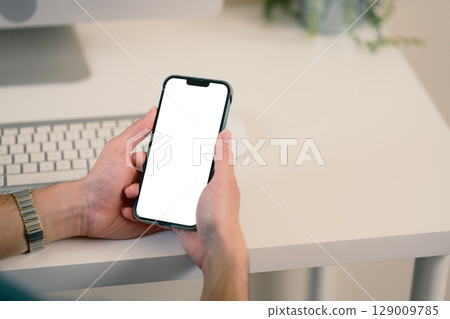 Close up of hands holding smartphone with blank white screen at a modern desk 129009785