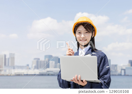 A young woman in work clothes using a laptop and pointing 129010108
