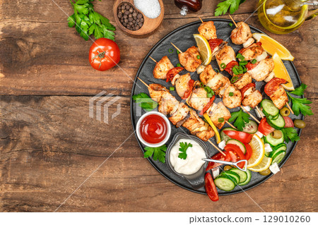 Top view of grilled chicken skewers with vegetables, sauces, and fresh salad on a rustic wooden table setting. top view. copy space Top view of grilled chicken skewers with vegetables, sauces, and fresh salad on a rustic wooden table setting. top view. copy space 129010260