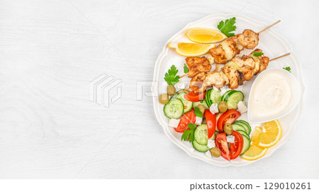 Chicken skewers served with fresh vegetable salad, lemon slices, and creamy sauce on a white plate over a bright wooden background. top view. copy space 129010261