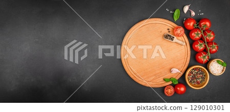 Round wooden cutting board with tomatoes, garlic, herbs, salt, and pepper on black kitchen background. Long banner format. top view. copy space. 129010301