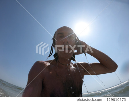 Handsome guy with long hair having fun at beach, sea waves splashing on his body 129010373