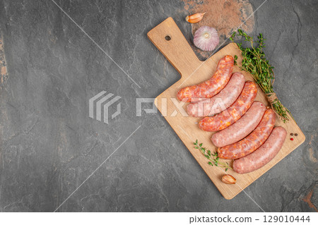 Raw pork sausages with garlic and herbs on a wooden board over a dark stone background. copy space, top view 129010444