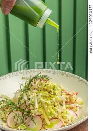 Fresh green dressing is delicately drizzled over a vibrant cabbage and radish salad, garnished with dill and flax seeds in a patterned bowl 129010475