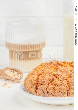 A freshly baked oatmeal cookie rests on a white plate beside a glass of layered coffee and scattered oats, creating a cozy breakfast scene 129010479