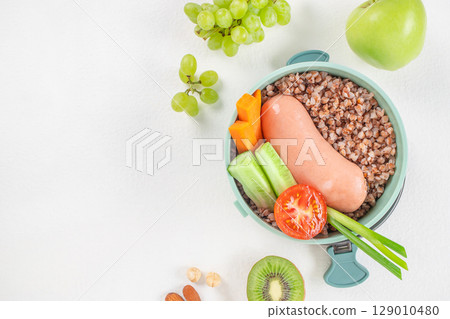 A balanced meal of buckwheat, sausage, fresh vegetables, and fruits is arranged in a lunchbox with a colorful, healthy presentation top view. copy space 129010480