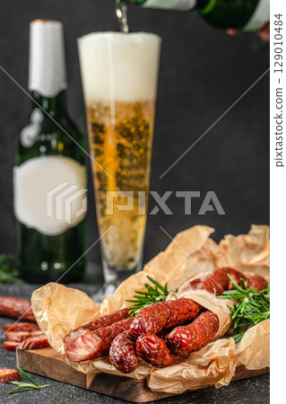 A tall glass of beer is being poured beside a rustic wooden board topped with smoked sausages and sprigs of fresh rosemary, creating a classic pairing of drink and charcuterie 129010484