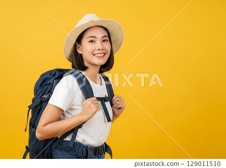 Adventurous Young Asian Woman with Backpack Smiles Cheerfully Against a Bold Yellow Background. Adventurous Young Asian Woman with Backpack Smiles Cheerfully Against a Bold Yellow Background. 129011510