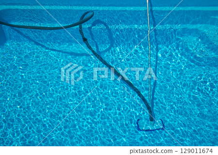 Blue hose and telescopic pole connected to a manual pool vacuum cleaner removing debris from the pool floor during outdoor summer maintenance. Blue hose and telescopic pole connected to a manual pool vacuum cleaner removing debris from the pool floor during outdoor summer maintenance. 129011674