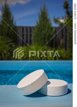 Two round chlorine tablets with blue specks resting on a poolside ledge in bright sunlight, used for water sanitation and pool care. 129011680