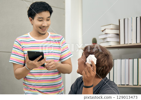 Two young men discussing real estate investment at home office, one smiling while holding tablet and other holding small house model, surrounded by bookshelves in modern indoor 129013473