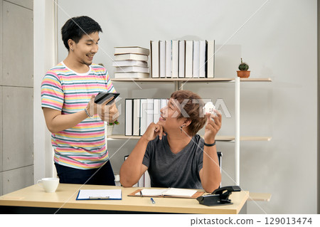 Smiling young men discussing property planning in home office, one seated holding small house model while other uses digital tablet, with documents and shelves in cozy workspace 129013474
