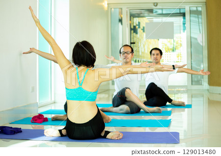 Group yoga session in bright indoor space with female instructor leading seated stretch pose, arms extended sideways, facing three adult men following her movements Group yoga session in bright indoor space with female instructor leading seated stretch pose, arms extended sideways, facing three adult men following her movements 129013480