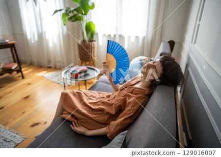 Tired woman use fan to relieve extreme heat on sofa soaked in sweat. Heatstroke risk, no AC 129016007