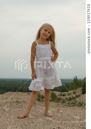 Happy young blond child girl in white dress standing on sand outdoors 129017419