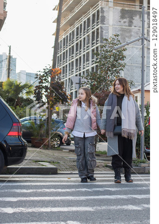 Cheerful mother and daughter hold each other's hand and cross the road at a crosswalk Cheerful mother and daughter hold each other's hand and cross the road at a crosswalk 129018497