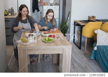 Concentrated daughter and her mother cooking in domestic kitchen together, family lifestyle portrait 129018863