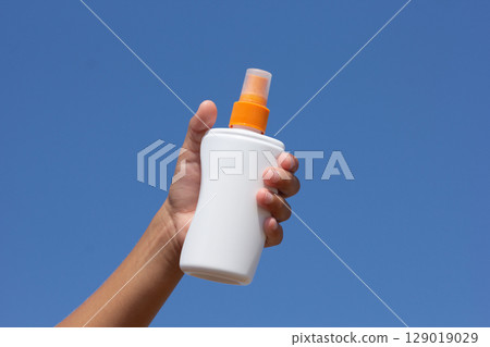 Sunscreen Spray Bottle in Childs Hand Against Blue Sky 129019029