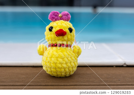 A cute, handmade crochet chick wearing a pink bow sits by a pool. Summertime vacation. A funky chicken toy against the blue water in a swimming pool. A cute, handmade crochet chick wearing a pink bow sits by a pool. Summertime vacation. A funky chicken toy against the blue water in a swimming pool. 129019283