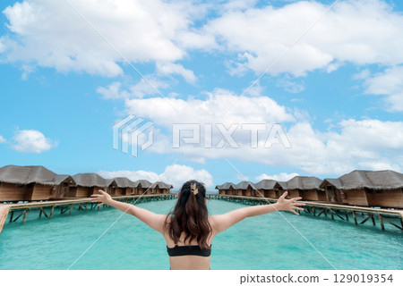 Stunning shot of a woman in a swimsuit with arms outstretched towards the beautiful ocean, framed by an idyllic overwater resort. Evokes luxury travel, relaxation, and dream vacations 129019354