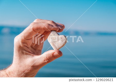 person holds heart shaped stone by water edge during a sunny day. closeup. person holds heart shaped stone by water edge during a sunny day. closeup. 129020621